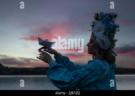 Young beautiful Ukrainian woman in a blue embroidered shirt, holding a paper boat made of white paper and looking forward with hope. Stock Photo