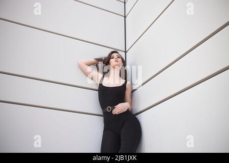 Young woman in black top and leggings leaning on white wall and looking at camera Stock Photo