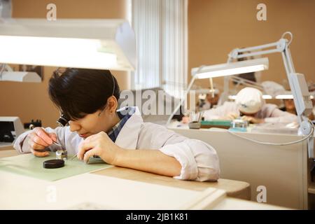 Serious concentrated female engineer sitting at table under illuminated lamp and assembling watch movements Stock Photo
