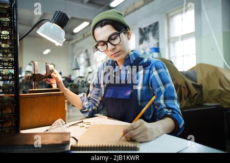 Serious busy hipster female worker in eyeglasses sitting at table and using stamping equipment while making holes into watch chips Stock Photo