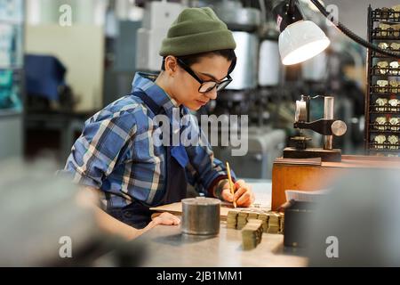 Serious concentrated female watch factory engineer in glasses sitting at desk and viewing notes while analyzing production of gears and cogs Stock Photo