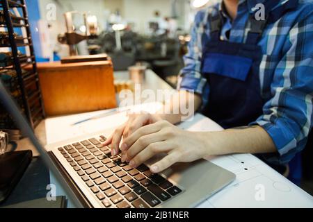 Close-up of unrecognizable woman engineer standing at desk with project plan and using laptop while searching for information on Internet Stock Photo
