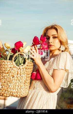 Portrait of young woman with long fair hair wearing white dress, floral earrings, standing near straw bag with bouquet. Stock Photo