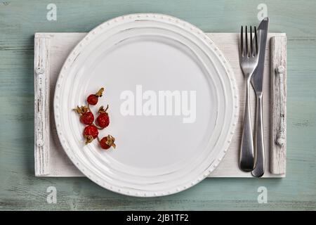 Handmade white ceramic plate with rose hips and cutlery on a white wooden tray. Mockup for displaying dishes Stock Photo