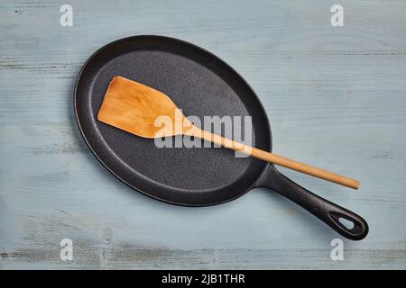 Oval cast iron frying pan with a wooden spatula on a blue wooden table. Menu template Stock Photo