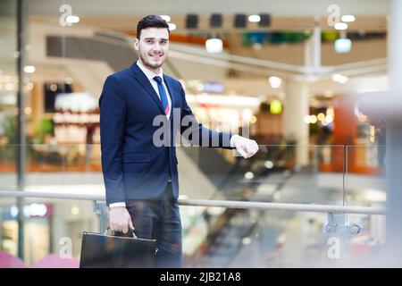 Smiling handsome young male property manager of shopping mall leaning on railing and holding briefcase while looking at camera Stock Photo