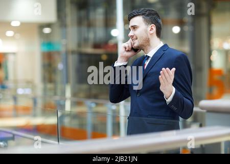 Content confident handsome young manager in formalwear standing in lobby and gesturing hand while solving work issues during phone conversation Stock Photo