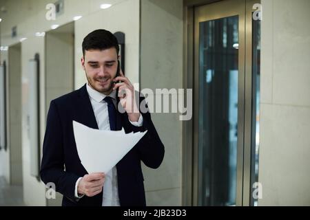 Cheerful confident handsome male manager in black suit walking by elevators and talking to business partner on phone while reading papers Stock Photo
