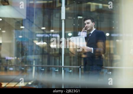Content confident multitasking young businessman in formalwear standing in lobby and discussing contract on phone, he working on move Stock Photo
