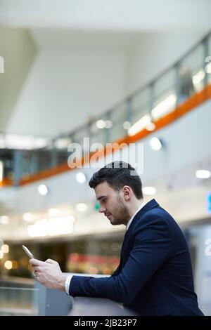Serious concentrated young man in formal suit leaning on railing and using gadget while wasting time in airport Stock Photo