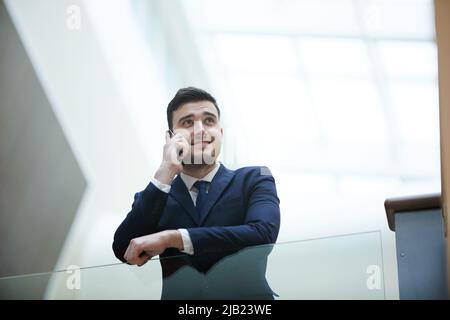 Smiling frowning handsome young businessman in suit leaning on glassy railing and communicating on cell phone while looking away Stock Photo