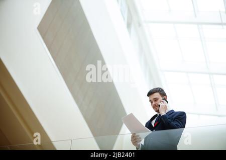 Below view of serious handsome young male manager in suit standing in modern lobby and analyzing papers while working with client on phone Stock Photo