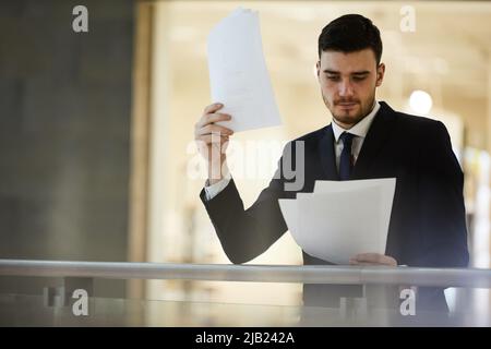 Serious thoughtful handsome young businessman in formalwear standing at railing in lobby and analyzing content of contract Stock Photo