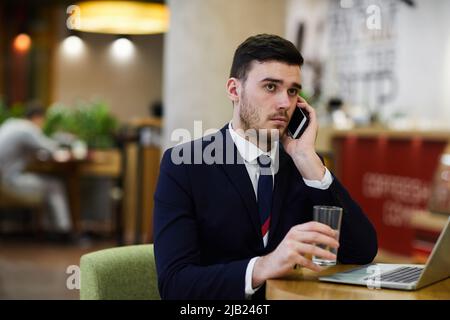 Serious pensive handsome young businessman with stubble sitting at table and drinking water while talking on phone Stock Photo