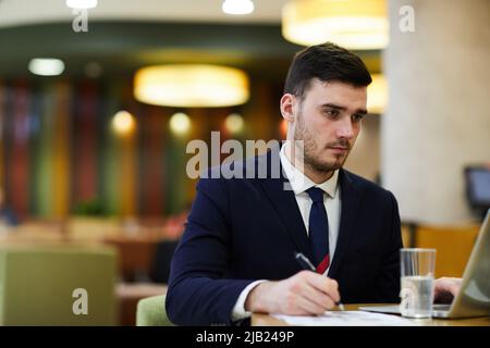 Serious concentrated handsome business expert in suit sitting at table in restaurant and editing information in papers while using laptop Stock Photo