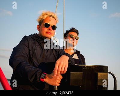 two persons on a sailboat, one person steering wheel sailing yacht other is standing near bow and looking forward at sunset, front view Stock Photo