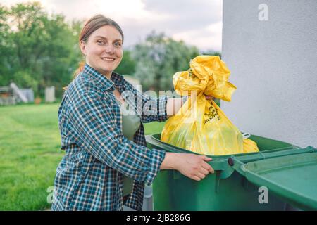 European 30s woman throwing garbage into the recycling bin in the backyard near the house. Separating recyclable garbage plastic  Stock Photo