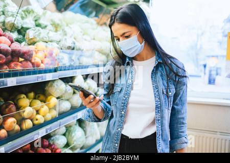 Brunette girl wearing protective mask uses smartphone while standing in supermarket shopping for food Stock Photo