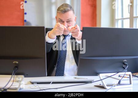 Businessman pointing with both hands at the camera viewed between computer monitors Stock Photo