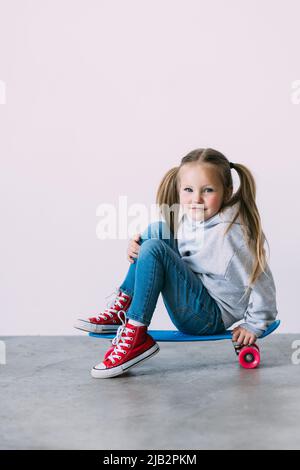 Smiling little girl in a summer dress fashion, sitting on a skateboard, and props up a head a hand, gazing at the camera with satisfaction and happine Stock Photo