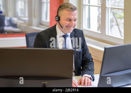 Smiling businessman wearing a headset seated at his desk having a teleconference call with a client Stock Photo