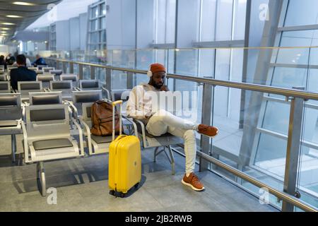Serious hipster black man sitting in airport terminal using laptop wear headphones listening music Stock Photo