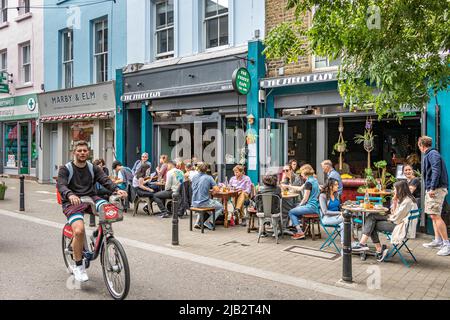 A man on a bike rides past people sitting outside eating and drinking on Exmouth Market, Clerkenwell ,London EC1 Stock Photo