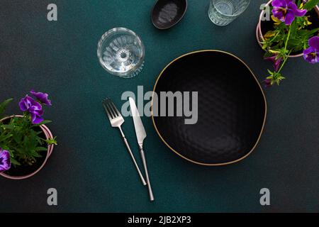 Table setting, empty black plate and cutlery on a green background, top view of the served table decorated with pansies flowers Stock Photo