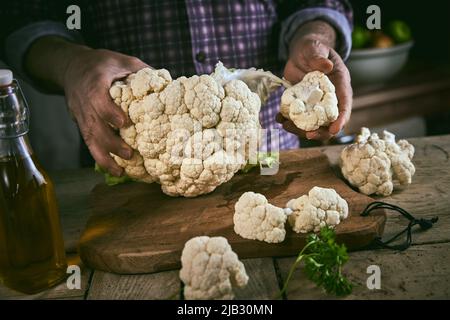 Anonymous male in checkered shirt dividing ripe cauliflower on smaller pieces on wooden cutting board near bottle of oil in rustic kitchen at home Stock Photo