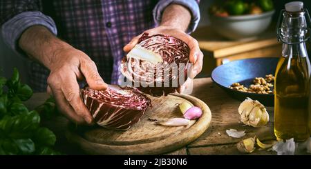 From above anonymous male in checkered shirt halving ripe red cabbage on round cutting board near cooking ingredients while preparing healthy dish in Stock Photo