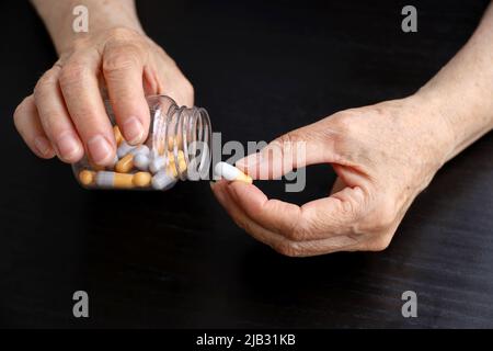 Wrinkled hands of elderly woman with pills on dark wooden table background. Medication in capsules, taking sedatives, antibiotics or vitamins Stock Photo