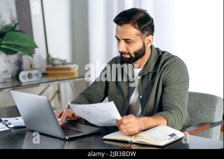 Focused successful indian or arabian businessman, trader, broker, company ceo, sits at a workplace in a modern office, studies statistics, looks at financial charts, works in a laptop, doing paperwork Stock Photo