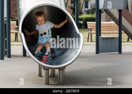 Boy in a white t-shirt blonde, riding on a metal slide tube on the playground. Stock Photo