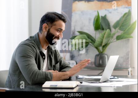Online conversation. Positive successful Arabian or Indian man, sitting in his modern office, talking with colleagues or client remotely via video call by laptop, discussing a project, smiles friendly Stock Photo