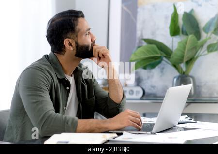 Pensive successful mixed race man, office worker, sitting in a modern office, distracted from work in a laptop, looking away thoughtfully, thinking about a new project, dreaming about vacation Stock Photo