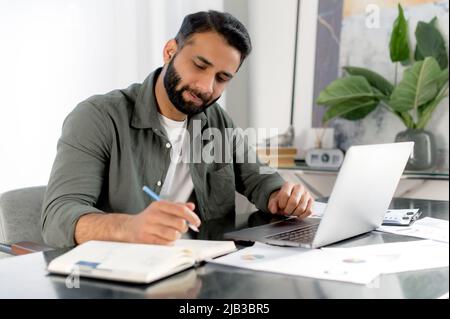 Smart busy focused man of Arabian or Indian nationality, successful company ceo, financial manager, sits at his desk in modern office, works with documents and laptop, analyzes, makes notes Stock Photo