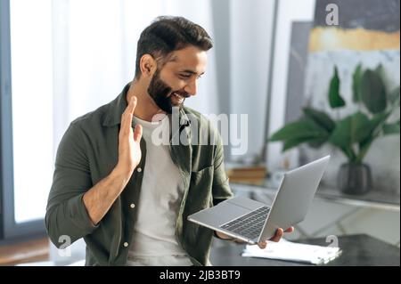 Successful arabian or indian man, holds and using laptop while stand in modern office, talking with coworker or client via video conference by laptop, conducts virtual meeting, smiling friendly Stock Photo