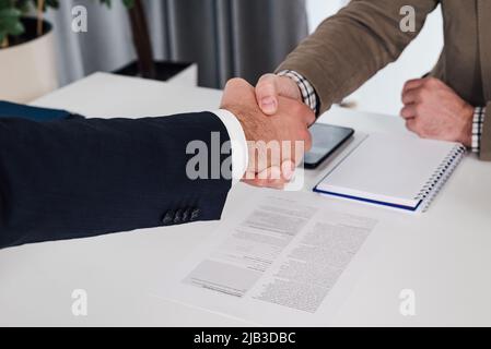 Male business partners shaking hands over contract. Professionals are sealing successful deal in meeting. They are sitting at desk in office. Stock Photo