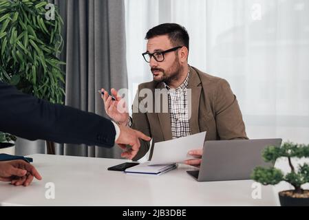 Confident young businessman discussing over contract with partner. Male executives planning deal during meeting at desk. They are in corporate office. Stock Photo