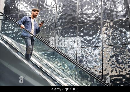 Front view of handsome young male going down by escalator. Stylish businessman holding smartphone, using, looking, scrolling, smiling. Concept of business and urban lifestyle. Stock Photo