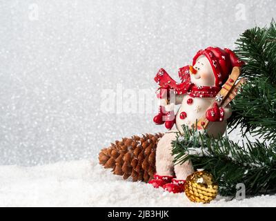 A snowman with skis sits on a pine cone. Christmas balls are spinning. Christmas and new year concept. Snow, pine, snowman, pine cone and decoration. Stock Photo