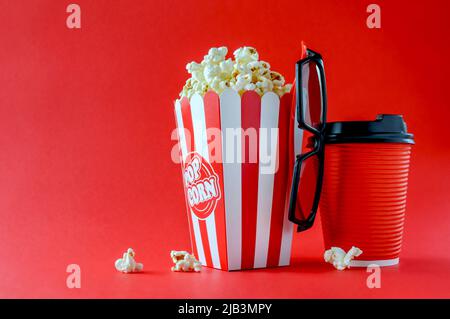 Popcorn in a striped cardboard box, 3d glasses, a paper glass with a drink on a red background, copy space. Movie Time. Stock Photo