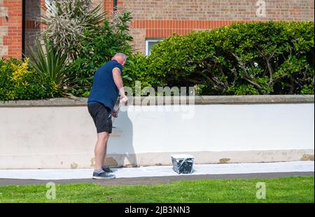 Man standing on a public pavement while painting an exterior garden wall in white, using a paint roller. Stock Photo
