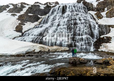 Dynjandi in Iceland looks especially massive next to two hikers standing on a rock in front of its cascade. Standing around 100 metres tall, it is the largest waterfall in the Westfjords, also known as Fjallfoss, and has been a protected natural monument since 1981. Patches of winter snow still cover large parts of the surrounding rocks in May. Stock Photo