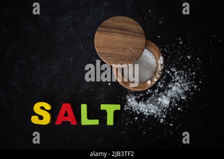 Top view of sea salt in wooden container on dark moody background with colored block lettering spelling the word salt.  Stock Photo