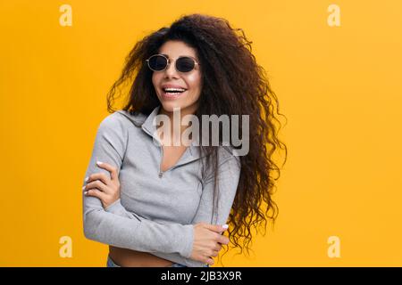 GOOD OFFER. Happy enjoyed pretty Latin female with afro in sunglasses crossing hands on chest, laughing, stay isolated over yellow background. Copy Stock Photo