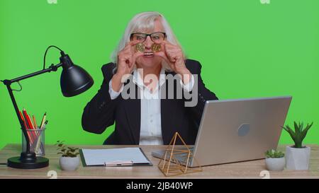 Elderly old business woman showing golden bitcoins. Achievement career wealth, cryptocurrency investment mining money income future technology. Senior woman freelancer fooling, making faces at office Stock Photo