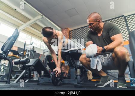 European young woman in a grey sports outfit lying on a gym bench leaning on a round weight, exercising her stomach muscles. Bald well-built man crunching near her holding a towel. High quality photo Stock Photo