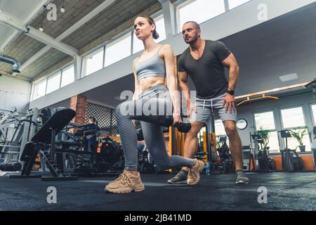 Gym time. Horizontal indoor wide shot of two caucasian people. Attractive pretty young woman doing exercise with weigths, good-looking hairless middle-aged man standing behind her. High quality photo Stock Photo