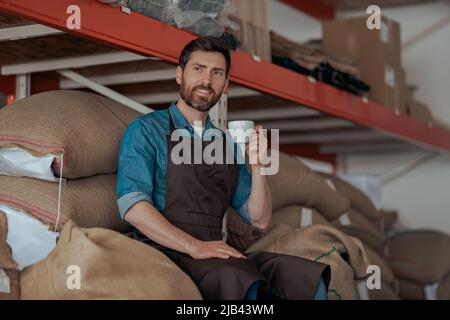 Business owner drinking coffee sitting on warehouse with bags of coffee beans  Stock Photo